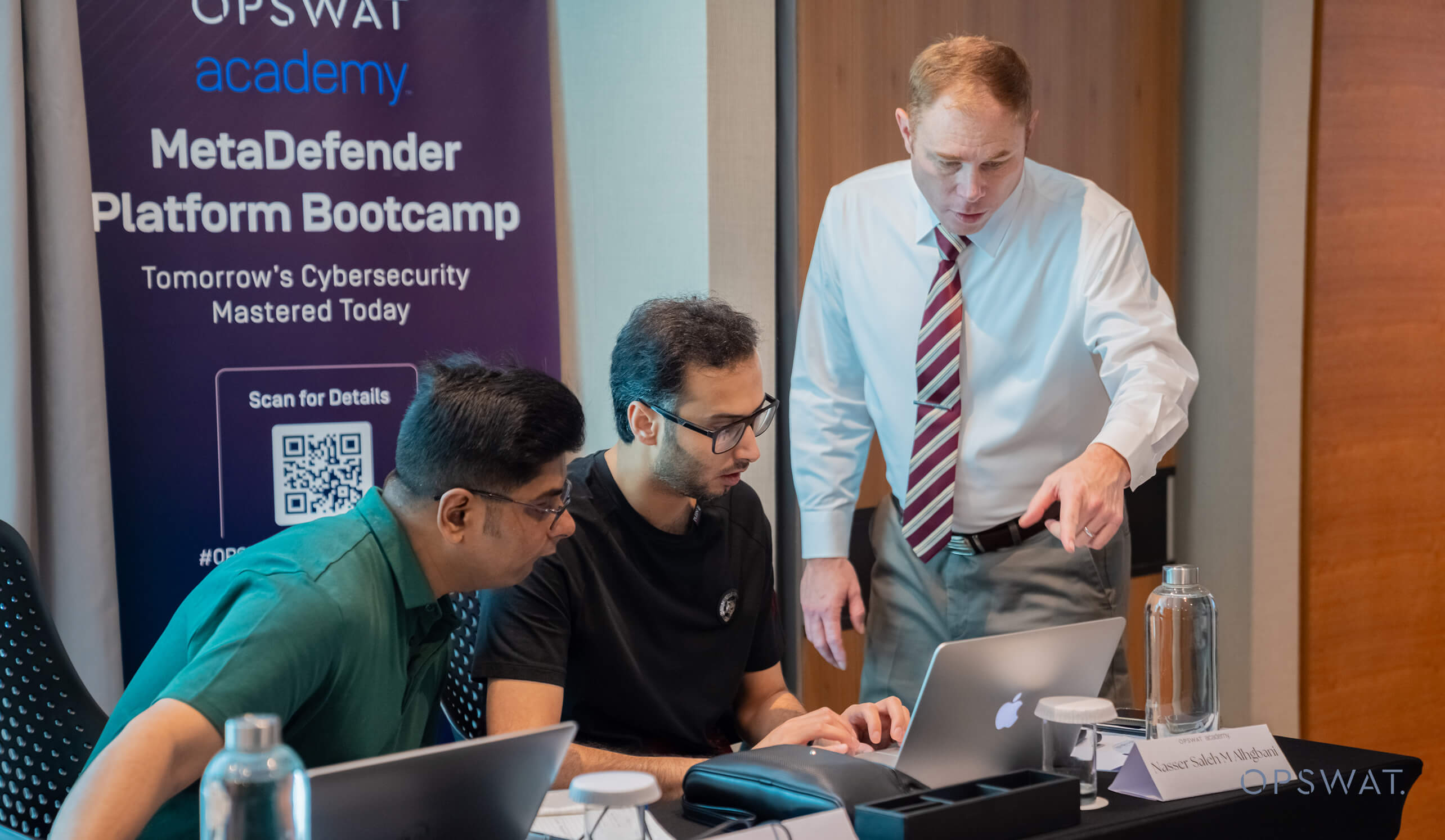 MetaDefender Platform Bootcamp Dubaiでラップトップで共同作業する3人。背景にはイベントのバナー。
