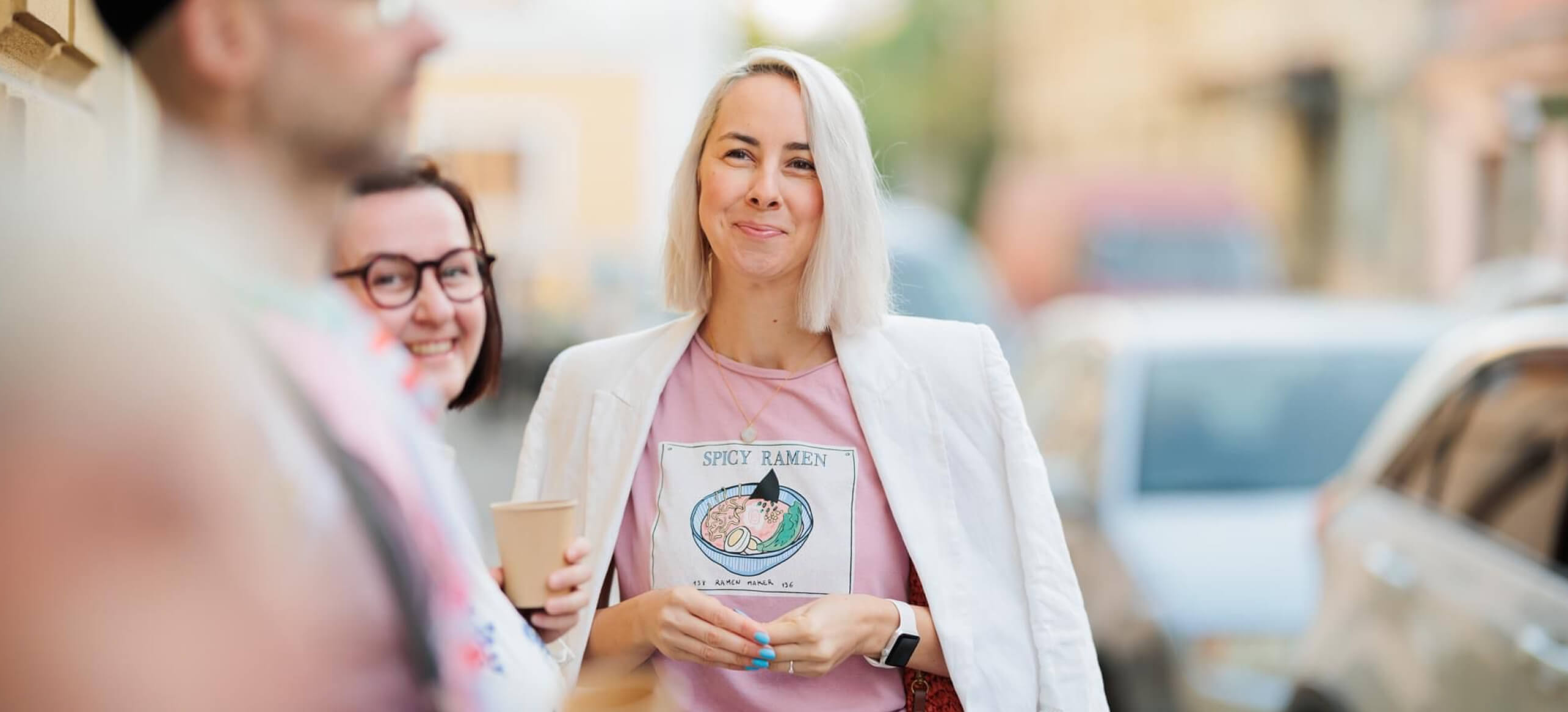 A smiling woman in a white blazer and a "Spicy Ramen" T-shirt enjoying a casual moment with two friends in an urban setting