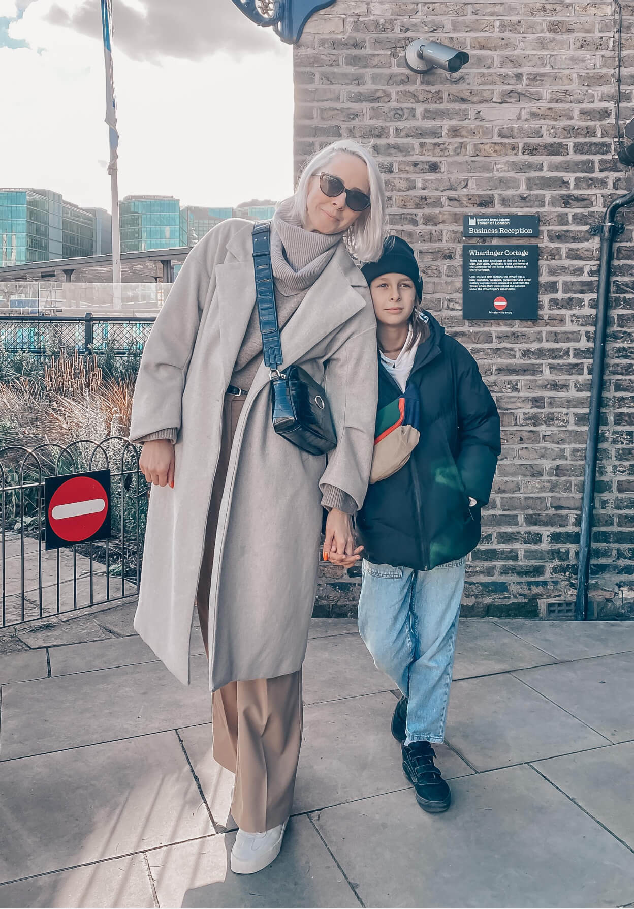 A woman in a long coat and sunglasses holds hands with a young person in casual wear, posing outdoors near a brick building