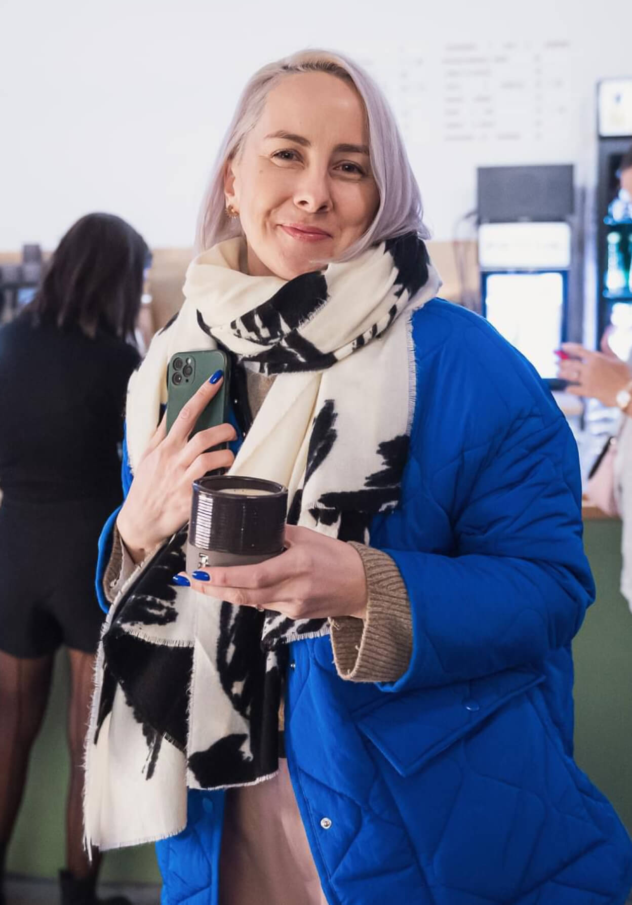 A woman in a blue puffer jacket and scarf holds a cup of coffee, smiling warmly in a café setting