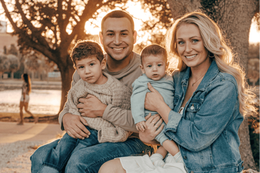 family portrait in a park setting with a Carlo, wife Brandee, and two young children smiling at the camera
