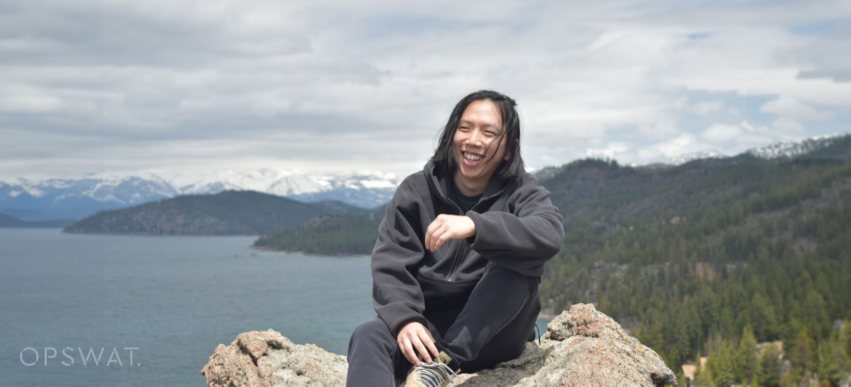 Randy Ying sitting on a rocky outcrop overlooking a lake with snow-covered mountains in the background