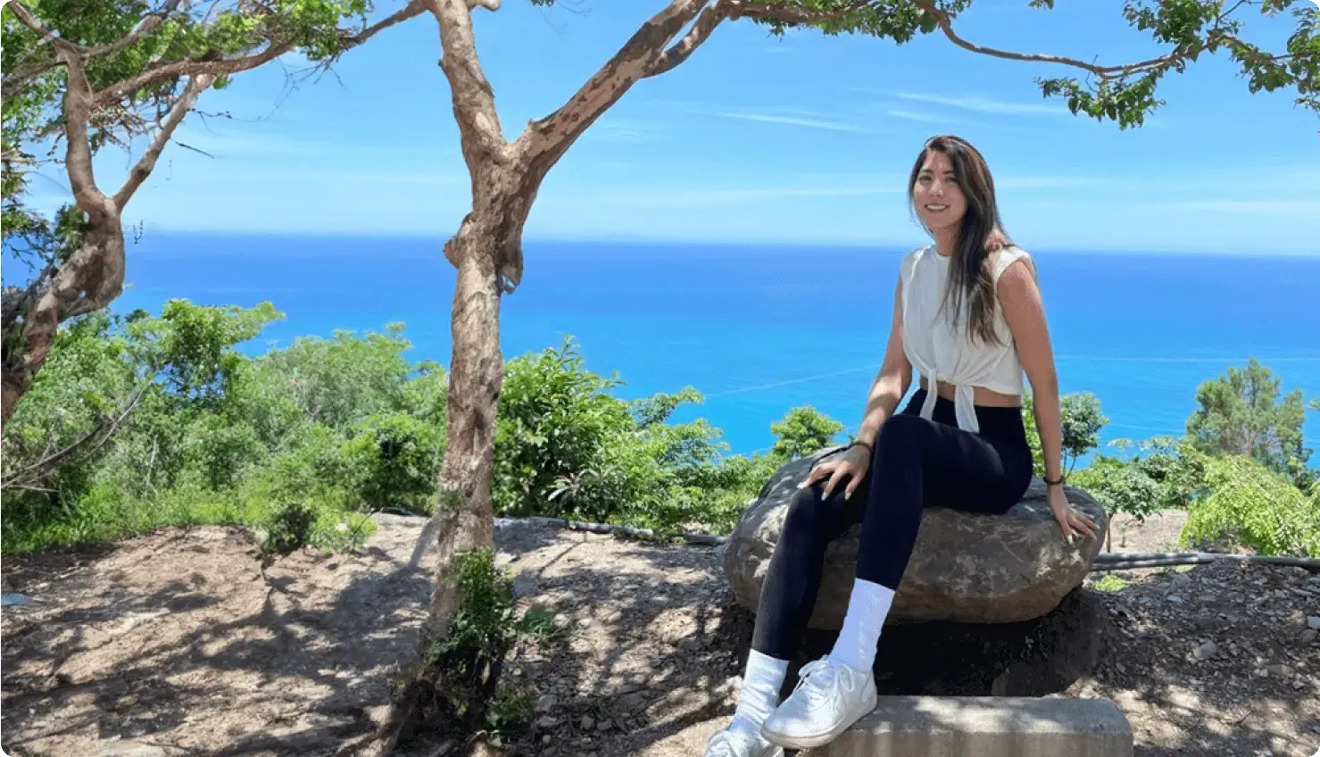 victoria huang sitting on a rock in front of a bright blue ocean