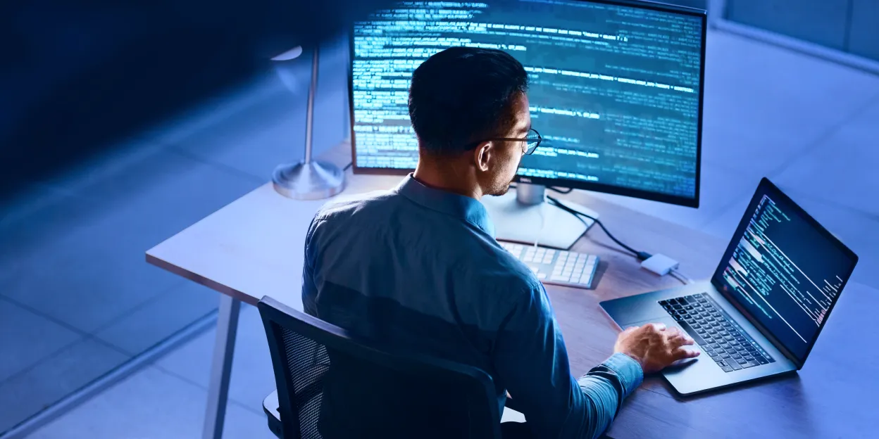 Person at desk with laptop and monitor displaying code, illustrating device posture check in a tech setting